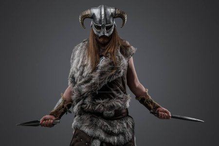 Foto de Tiro de vándalo nórdico con piel y casco sosteniendo dos dagas sobre fondo gris. - Imagen libre de derechos