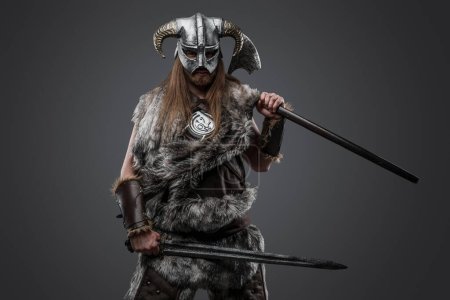 Photo for Shot of scandinavian warrior dressed in fur holding sword and axe against grey background. - Royalty Free Image