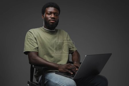 Téléchargez les photos : Studio shot de mec noir avec ordinateur portable assis sur la chaise sur fond gris. - en image libre de droit