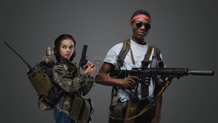 Téléchargez les photos : Prise de vue de petite fille avec arme de poing et homme noir avec fusil sur fond gris. - en image libre de droit