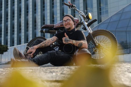 Foto de Tiro de balancín elegante sonriente con moto mostrando pulgares hacia arriba signo al aire libre. - Imagen libre de derechos