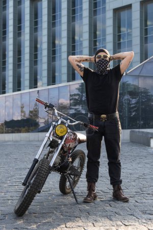 Téléchargez les photos : Portrait de motard urbain tatoué avec bandana et moto posant à l'extérieur en ville. - en image libre de droit