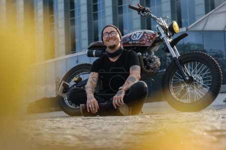 Téléchargez les photos : Prise de vue de rocker élégant souriant avec moto regardant la caméra à l'extérieur. - en image libre de droit