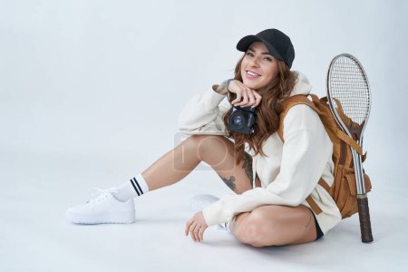Photo for Sports gear and lifestyle. Beautiful sportswoman wearing white hoody with baseball cap poses in studio with photo camera and racquet. - Royalty Free Image