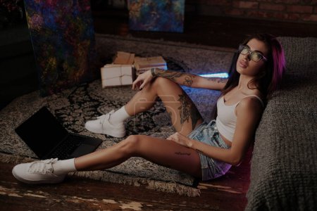 Foto de Retrato atmosférico de una chica glamurosa vestida con gafas y con portátil sentado en el suelo alrededor de un rayo de neón. - Imagen libre de derechos