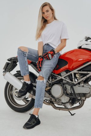 Téléchargez les photos : Plan studio de femme active aux longs poils blonds chevauchant une moto rouge sur fond blanc. - en image libre de droit