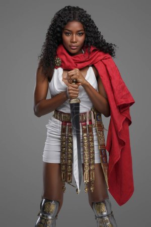 Téléchargez les photos : Modèle afro-américain exquis en tunique blanche avec une belle cape rouge ornée de tombes de ceinture tout en posant avec confiance avec gladius épée sur fond gris - en image libre de droit