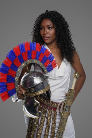 Téléchargez les photos : Femme afro-américaine élégante porte une robe blanche de style romain avec une ceinture ornée et des cretons, tenant un casque de gladius et de centurion romain, sur un fond gris - en image libre de droit