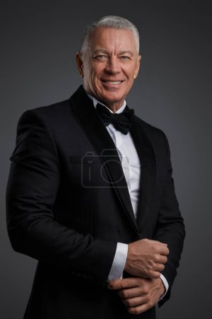 Téléchargez les photos : Elégant homme âgé, vêtu d'un costume noir et d'un nœud papillon, pose avec confiance avec un sourire charmant pour une séance photo sur un fond gris - en image libre de droit