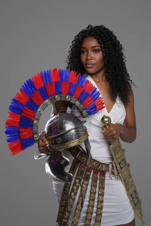 Téléchargez les photos : Femme afro-américaine élégante porte une robe blanche de style romain avec une ceinture ornée et des cretons, tenant un casque de gladius et de centurion romain, sur un fond gris - en image libre de droit