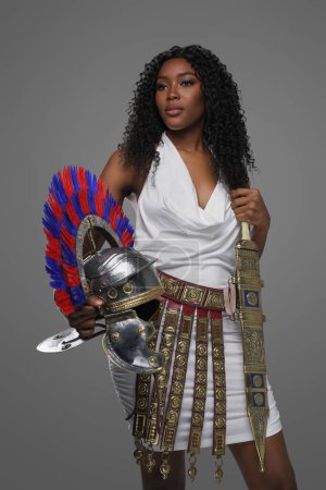Foto de Elegante mujer afroamericana lleva un vestido blanco de estilo romano con un cinturón adornado y chicharrones, sosteniendo un casco de gladius y centurión romano, sobre un fondo gris - Imagen libre de derechos