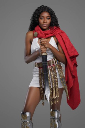 Foto de Modelo afro-americano exquisito en túnica blanca con una hermosa capa roja tumbas de cinturón adornado mientras posa con confianza con espada de gladius sobre fondo gris - Imagen libre de derechos