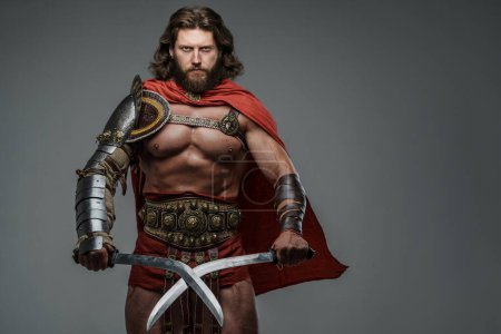 Foto de Un fiero e imponente gladiador de pelo largo y barba, con armadura ligera y de pie con dos espadas. Este guerrero exuda fuerza y poder sobre un fondo gris neutro - Imagen libre de derechos