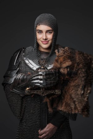 Téléchargez les photos : Chevalier féminin confiant en armure médiévale, souriant largement et posant avec une épée à la main tandis qu'il était drapé de fourrure, sur un fond gris - en image libre de droit