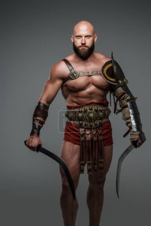 Foto de Gladiador calvo y barbudo posa de pie con dos espadas mientras usa armadura ligera. Este temible e imponente guerrero emana poder y fuerza sobre un fondo gris neutro - Imagen libre de derechos