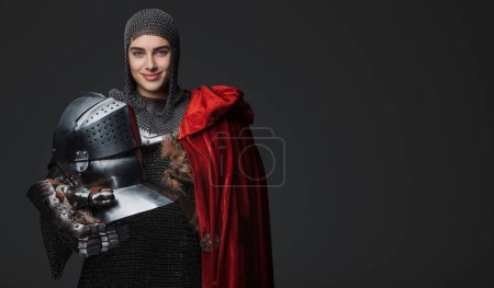 Foto de Caballero femenino confiado en armadura medieval, sonriendo ampliamente y posando con un casco, cubierto con una capa roja, sobre un fondo gris - Imagen libre de derechos