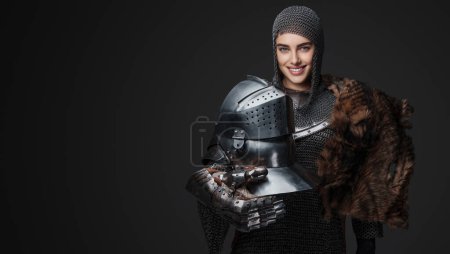 Foto de Hermosa mujer caballero con armadura medieval, sonriendo amplia y confiadamente posando con una piel cubierta sobre un hombro y un casco en la mano, sobre un fondo gris - Imagen libre de derechos