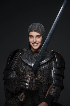 Photo for Beautiful woman is dressed in Knightly armor, is smiling wide, and confidently posing with a sword in-hand, and draped in fur. The photograph is set against a grey background - Royalty Free Image