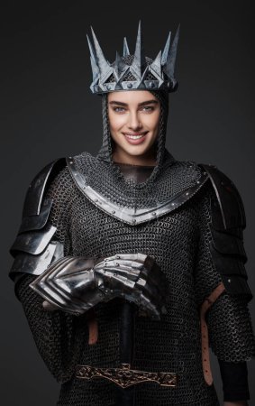 Photo for Beautiful queen knight in medieval armor and steel crown exudes joy and confidence as she wields her sword. Her broad smile and relaxed pose highlight the spirit of triumph and pride - Royalty Free Image