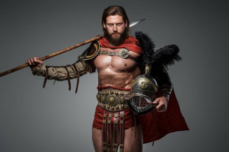 Foto de Un gladiador barbudo con el pelo largo está parado sobre un fondo gris en armadura ligera y una capa roja, sosteniendo una lanza y un casco con plumas - Imagen libre de derechos
