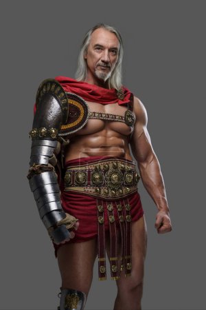Photo for Aged but still mighty, this muscular gladiator with long grey hair and beard stands proudly in lightweight armor against a grey background - Royalty Free Image