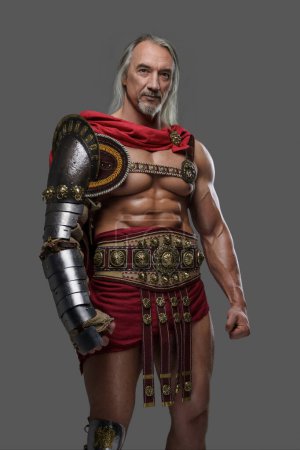 Foto de Gladiador maduro y musculoso con una elegante barba gris y cerraduras de plata que fluyen dons armadura ligera posando sobre fondo gris - Imagen libre de derechos