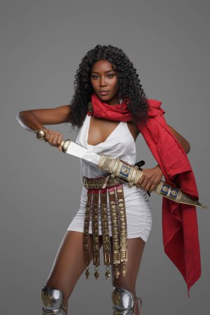 Photo for Stunning dark-skinned beauty dons a white Greek tunic with a striking red cape, intricately decorated belt and greaves and confidently wields richly adorned gladius sword on grey background - Royalty Free Image