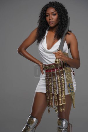 Photo for Stunning afro-American beauty with curly hair and adorned in a white Greek tunic, embellished with ornate belt and leg greave, poses with a gladius on a grey background - Royalty Free Image
