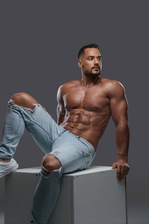 Remarkably attractive male model with a muscular physique poses topless in ripped jeans on a white cube against a grey backdrop, exuding sexiness and ruggedness