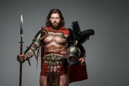 Téléchargez les photos : Un gladiateur barbu aux cheveux longs se dresse sur un fond gris en armure légère et un manteau rouge, tenant une lance et un casque avec des plumes - en image libre de droit