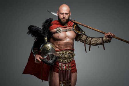 Gladiador calvo seguro con armadura ligera y capa roja, sosteniendo una lanza y casco de gladiador con plumas, mirando hacia abajo la cámara sobre un fondo gris neutro