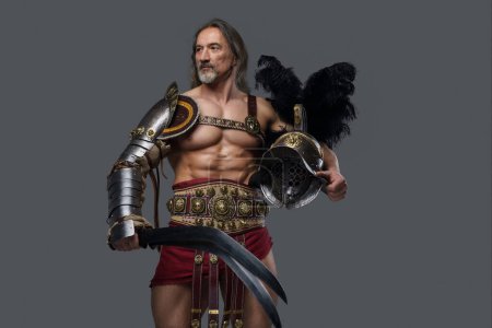 Photo for Imposing gladiator of mature age with a stylish silver beard and flowing grey hair dons lightweight armor, wielding a gladius and holds helmet as he stands proud against a neutral grey backdrop - Royalty Free Image