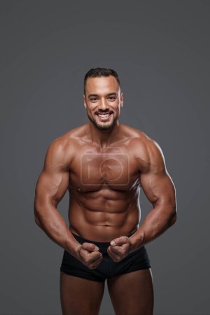 Photo for Breathtakingly handsome male model with a chiseled physique flashes a charming smile as he poses in black briefs against grey background, emanating charisma and sex appeal - Royalty Free Image