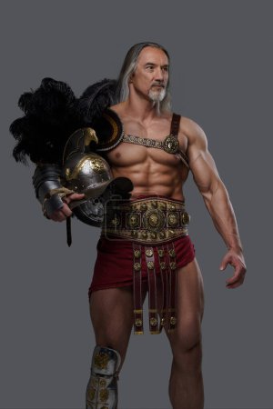 Photo for Mature muscular gladiator with a stylish grey beard and flowing silver locks dons lightweight armor, holding a gladiator helmet with a feathered crest while posing against a neutral grey backdrop - Royalty Free Image