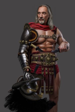 Téléchargez les photos : Gladiateur distingué avec une barbe argentée élégante et des serrures pulpeuses porte une armure légère, tenant un casque de gladiateur avec panache à plumes alors qu'il se tient avec confiance sur fond gris - en image libre de droit