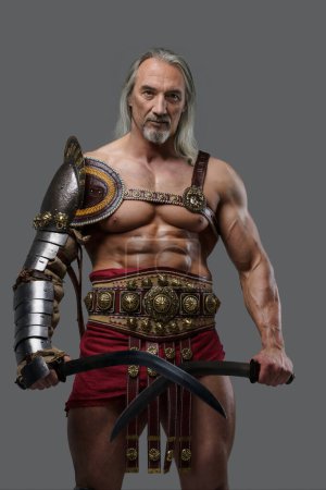 Photo for Distinguished elder gladiator exudes power and strength in elegant, lightweight armor, wielding two gladius swords with confidence against a grey background - Royalty Free Image