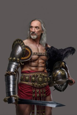 Photo for This regal, aged gladiator exudes strength and dignity in sleek, lightweight armor, holding a gladius sword and helmet against a grey background - Royalty Free Image