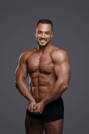 Photo for Breathtakingly handsome male model with a chiseled physique flashes a charming smile as he poses in black briefs against grey background, emanating charisma and sex appeal - Royalty Free Image
