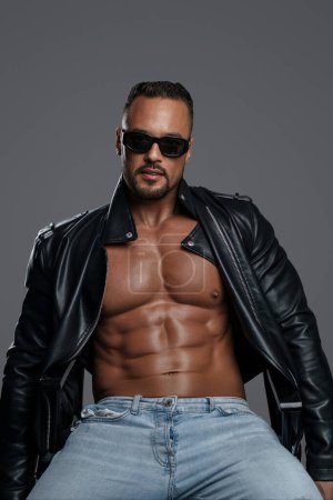 Photo for Attractive male model with a muscular shirtless torso poses in ripped jeans, a black leather jacket, and dark sunglasses while seated on a studio chair against a grey backdrop - Royalty Free Image