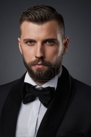Téléchargez les photos : Portrait en gros plan d'un homme élégant et séduisant à la barbe soignée, vêtu d'un costume noir haut de gamme et d'un nœud papillon, posant en toute confiance sur un fond gris - en image libre de droit