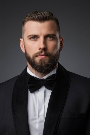 Close-up portrait of a stylish and attractive man with a well-groomed beard, dressed in a high-end black suit and bow tie, posing confidently on a gray background