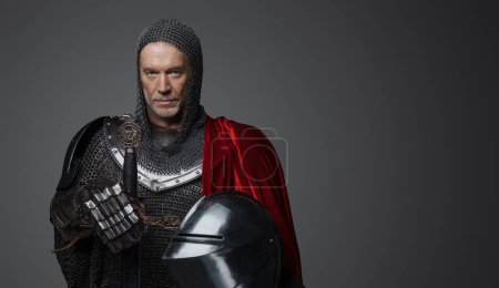 Photo for Medieval-style portrait of a charismatic king with a gray beard in chainmail armor with steel plates, holding a sword and posed against gray - Royalty Free Image