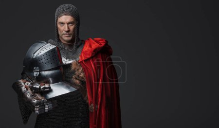 Photo for This portrait encaptures a medieval warrior in armor with a red cape draped across one shoulder. The powerful knight stands with a commanding presence, gripping his helmet with one hand - Royalty Free Image