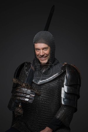 Téléchargez les photos : Chevalier mature en armure lourde, portant un large sourire et brandissant son épée avec confiance, pose sur un fond gris - en image libre de droit