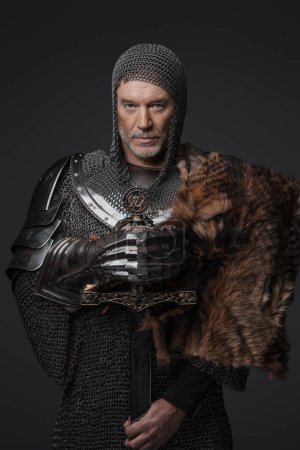 Foto de Retrato de estilo medieval de un rey carismático con una barba gris en armadura de malla de malla con placas de acero, sosteniendo una espada y posando contra gris - Imagen libre de derechos