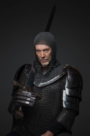 Photo for Confident portrait of a mature king with a gray beard, wearing heavy armor, holding a sword against a gray background - Royalty Free Image