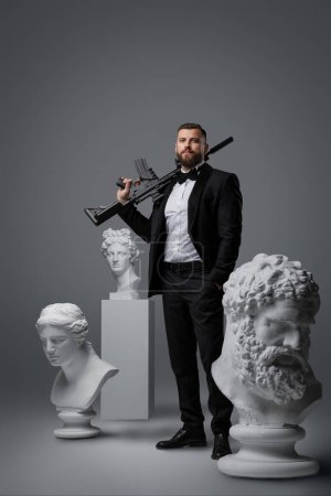 Photo for Stylish and enigmatic man in a black suit and bow tie holding a gun, embodying a secret agent or spy, posing with confidence in front of antique statues on a gray background - Royalty Free Image