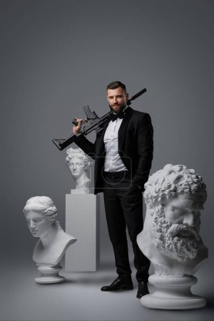 Photo for Stylish man with a well-groomed beard dressed in a luxurious black suit and bow tie, holding an automatic rifle striking a confident pose among three antique statues against a grey background - Royalty Free Image