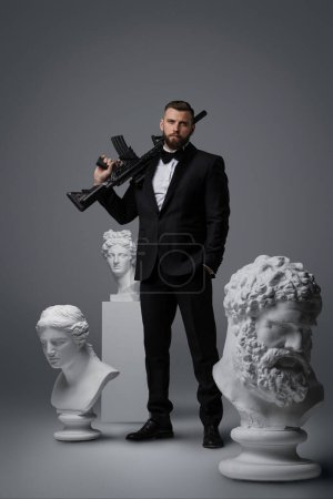 Photo for Stylish man with a well-groomed beard dressed in a luxurious black suit and bow tie, holding an automatic rifle striking a confident pose among three antique statues against a grey background - Royalty Free Image
