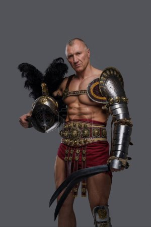 Photo for Battle-worn muscular gladiator of mature age with a rugged face dons intricately designed lightweight armor, wielding a gladius and donning a feathered helmet against grey background - Royalty Free Image